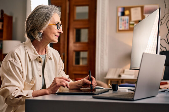 A beautiful older woman works in a home office using a graphics tablet .Creative profession, remote work, learning.