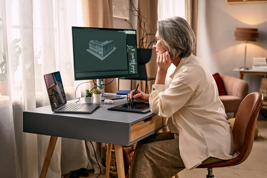 Mature woman working at a home office desk using a pen tablet and dual screens showing 3d architectural model. Remote professional work at home.