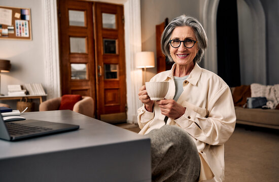 Smiling older woman with gray hair and glasses holding a mug in a cozy, well-lit living room office.