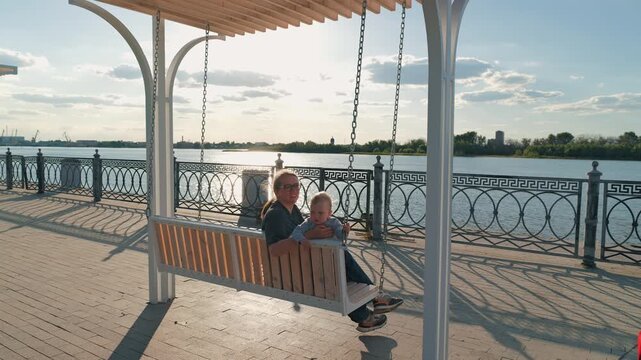 Happy mother with son swinging on wooden bench on riverfront promenade. Family rests together outdoors by water during golden hour sunset. Warm, serene moment with long shadows
