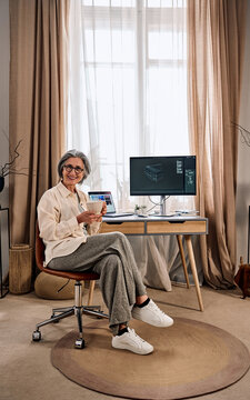 Smiling older woman sitting in a cozy home office, holding a mug, with desk, laptop and monitor, soft natural light from large window.