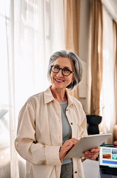 Smiling confident middle-aged woman holding a digital tablet, looking at the camera in a modern home office setting, vertical portrait