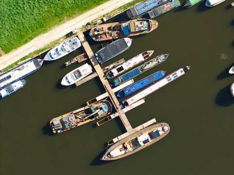 Aerial view of various boats and narrowboats moored at a wooden pier in a marina with green grass and a path alongside the dark water Rawcliffe Bridge, England, United Kingdom.