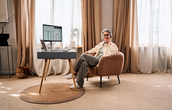 Mature woman relaxing in a comfortable armchair in a cozy beige-toned living room at home, sitting by a desk, working from home