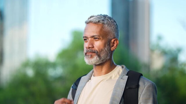Confident mature Brazilian businessman with backpack walking relaxed on urban sidewalk with hand on chin in city