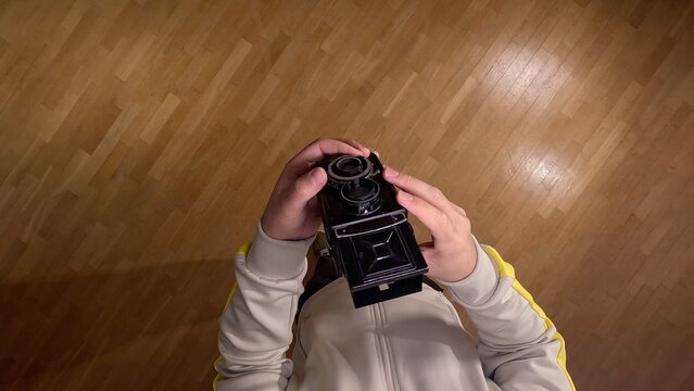 Users hands holding a black vintage twin lens reflex camera, adjusting settings while standing on a warm wooden parquet floor, capturing a moment of analog photography