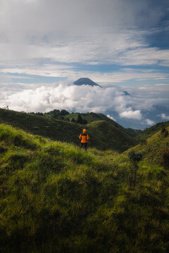 Aerial view of Gunung Prau with a hiker in an orange jacket looking out over a sea of clouds and distant volcanic peaks Mount Prau, Central Java, Indonesia.