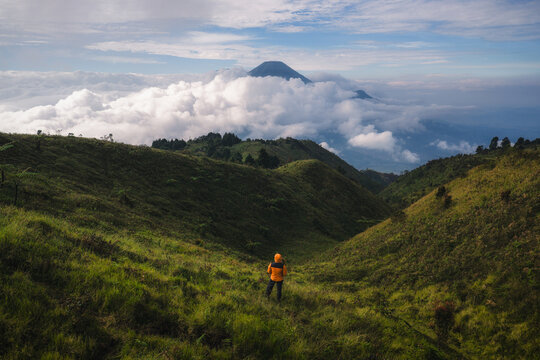 Aerial view of a hiker in an orange jacket standing on the grassy slopes of Gunung Prau overlooking a sea of clouds and distant volcanic peaks Mount Prau, Central Java, Indonesia.