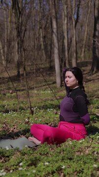 Vertical video. Peaceful woman in workout attire sits in yoga pose on grass stretching spine. Serene female in training clothes sits on forest lawn gently pulling arms backward to stretch upper back