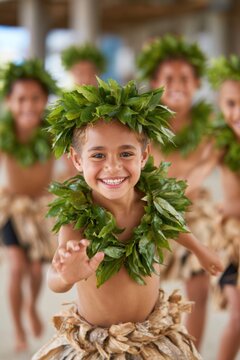 Boys performing a traditional cultural dance wearing leaf crowns and grass skirts, smiling and dancing joyfully in a coastal outdoor setting
