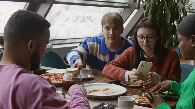 Young woman with smartphone taking pictures of diverse friends putting pizza slices on their plates while spending time together in cafe