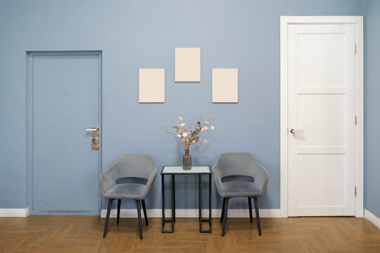 Minimalist waiting room with modern gray armchairs, side table holding a vase of dried flowers, three blank frames on a soothing blue wall and warm wooden flooring