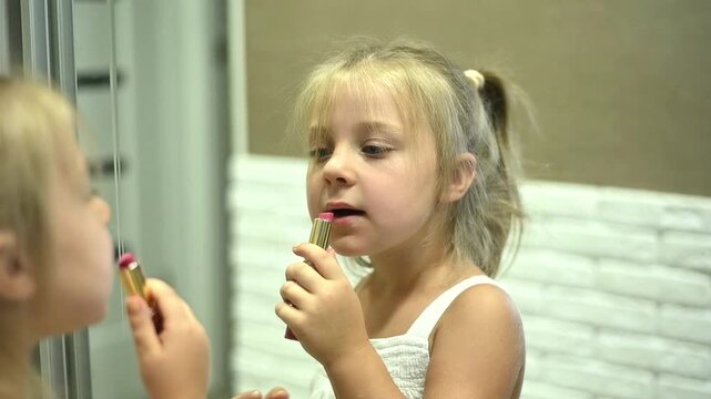 Little girl applying lipstick while looking in mirror, small child holding lipstick and carefully coloring lips in playful imitation of adult makeup. moment with pomade. childhood lifestyle, beauty