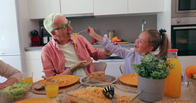 Playful little granddaughter feeding her senior grandfather spaghetti with a fork during a happy family lunch at home. Loving relationship between generations enjoying a meal together in the kitchen