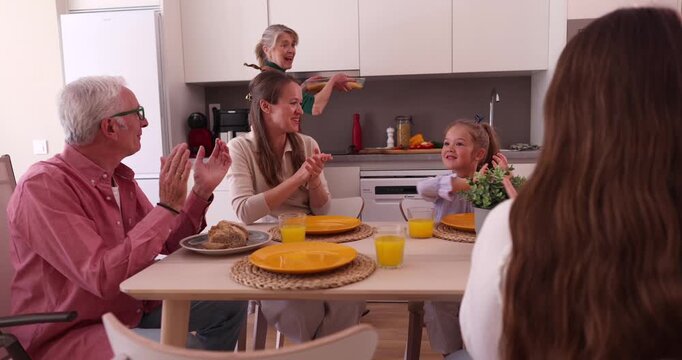 Happy three generation family clapping hands and celebrating as the senior grandmother brings a freshly baked dish to the dining table for a joyful holiday meal together in the kitchen