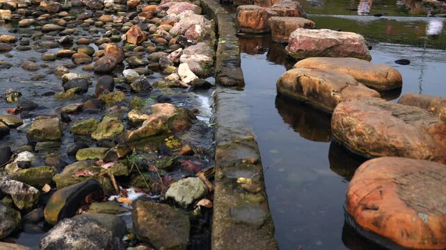 Tranquil city park river stream with stepping stones and flowing water over concrete dam 4K.a peaceful urban river landscape, featuring large natural stepping stones, a small stone dam with cascading 