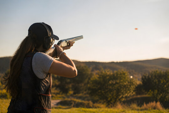 Woman aiming shotgun at clay target during outdoor shooting practice at sunset