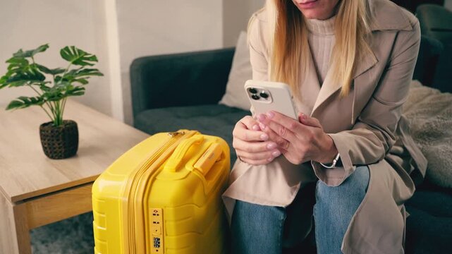 Young businesswoman with her yellow suitcase sitting on the sofa in a modern apartment, booking a hotel or ordering a taxi online with her smartphone before a business trip