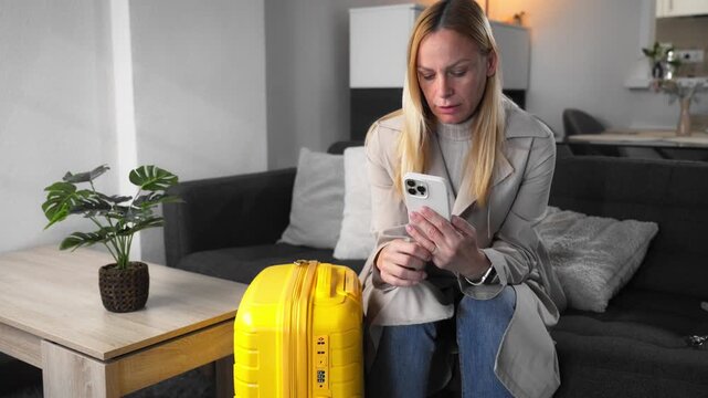 Woman sit in room dressed with suitcase, scrolling on phone,touch wedding ring.Concept of emotional decision-making, relationship uncertainty, life-changing choices before leaving or start new chapter
