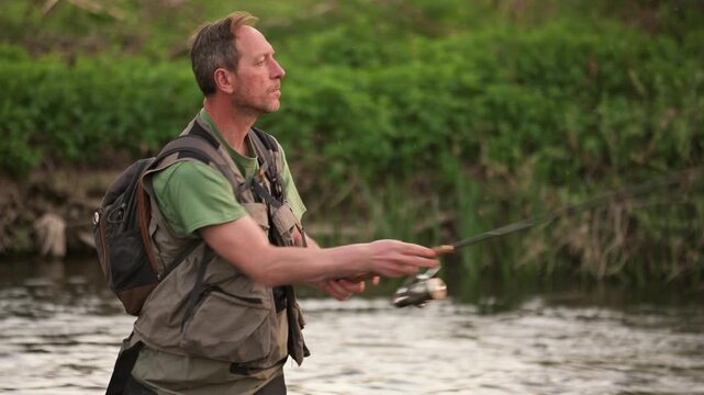 Medium shot of fisherman casting rod while standing in shallow river water. Focused angler enjoying peaceful fishing session in natural surroundings. High quality 4k footage