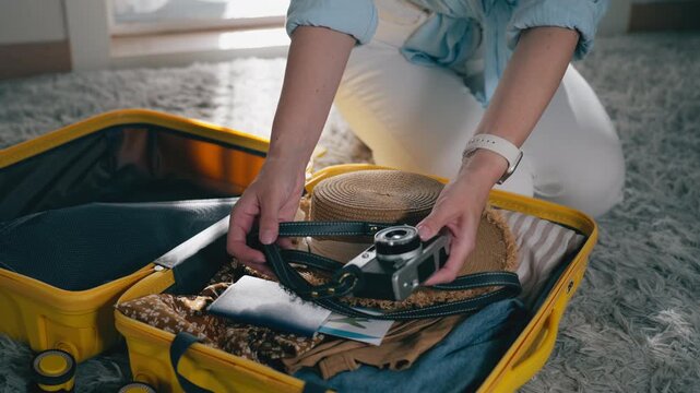 Close-up of hand placing vintage film camera into an open yellow suitcase filled with travel essentials. Concept of summer vacation preparation, travel packing,anticipation of holiday adventure abroad