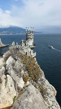 Swallow's nest castle perched on gagarin rock cliff above the dark black sea, dramatic neo-gothic silhouette, boat below and distant mountains under a partly cloudy sky