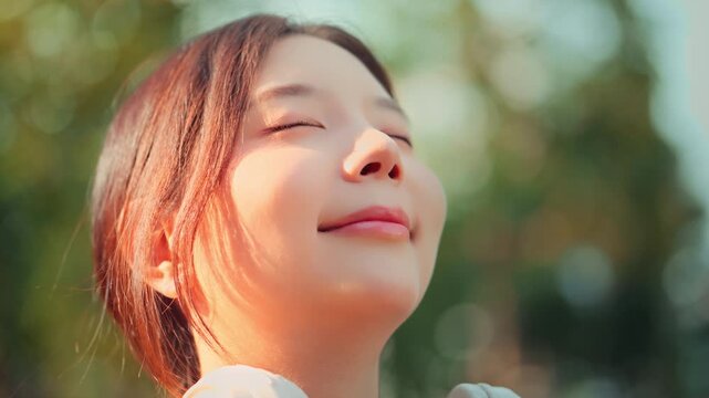 Close up of happy young Asian woman eyes closed smiling in natural sunlight at park. Cinematic portrait for mindfulness, wellness, skincare, and mental health lifestyle concept. Beautiful female face 
