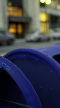A blue mailbox is positioned on a city street. Cars are parked along the road. Buildings in the background are lit up as evening falls.