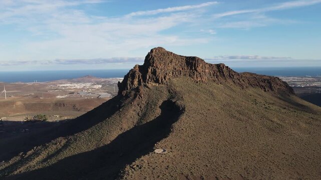 Aerial view circling the volcanic peak Roque Aguayro on Gran Canaria island. Scenic landscape with the coastline, a town, and a wind farm in the background