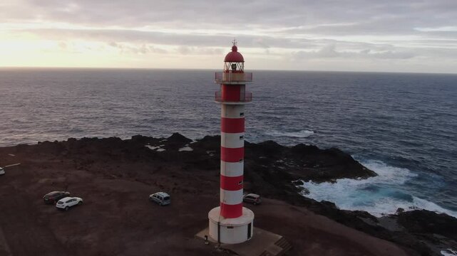 Aerial view of the iconic Sardina del Norte lighthouse standing on a rocky coast in Gran Canaria, with waves crashing against the shore at sunset