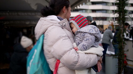 Fototapeta premium Mother carrying baby in crowded winter street, dressed warmly in coat and hat, affectionate everyday family moment showing warmth, protection, and love