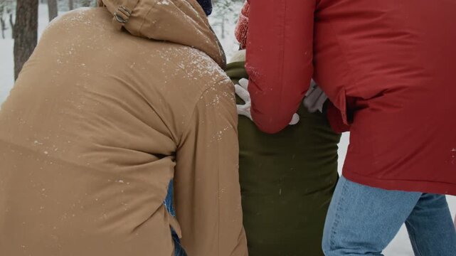 Two friends pushing young man who sliding down slope on red sled raising his hands up in snowy forest on winter day
