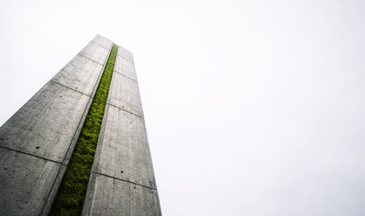 Fototapeta premium Low angle view of a tall concrete brutalist pillar with a vertical green moss garden strip, concept of sustainable architecture and urban greening