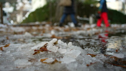 Naklejka premium Melting snow on city sidewalk with fallen leaves and reflections, subtle sign of seasonal change as people walk in background, early winter thaw and urban transition
