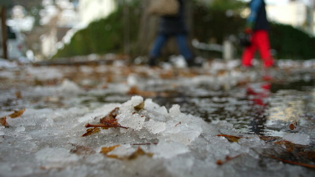 Melting snow on city sidewalk with fallen leaves and reflections, subtle sign of seasonal change as people walk in background, early winter thaw and urban transition