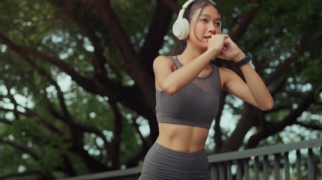 Active young Asian woman in sportswear wearing white headphones and smartwatch performing squats at an outdoor park during golden hour. Concept of modern fitness, healthy lifestyle, wellness