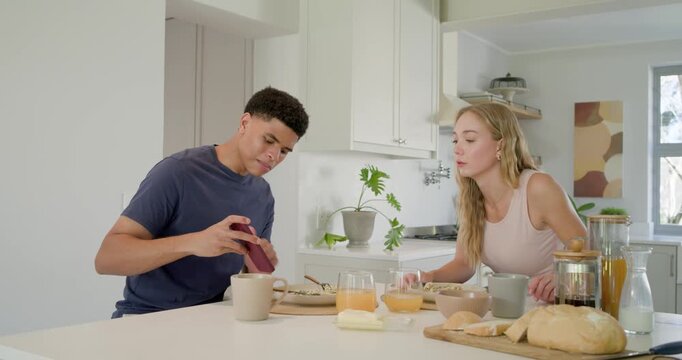 Diverse couple sitting at kitchen island, man picking maroon bottle, trying to open, woman watching