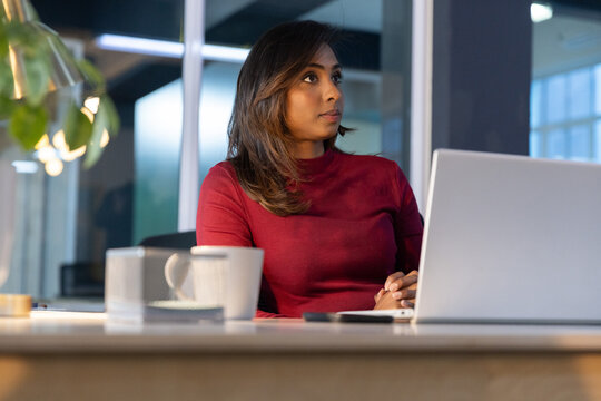 Indian adult woman wearing maroon long-sleeve top typing on laptop holding mug at desk