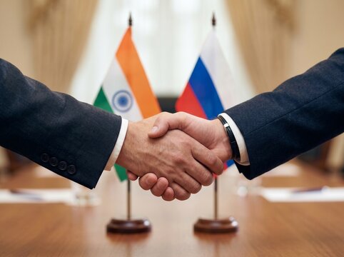Two businessmen in suits shaking hands firmly with Indian and Russian flags displayed in the background symbolizing a significant diplomatic or trade agreement being finalized and confirmed.