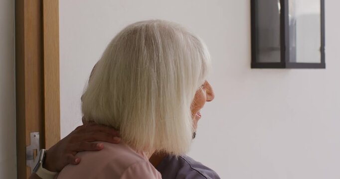 Adult woman in gray scrubs arriving at doorway, hugging senior woman, giving visit update