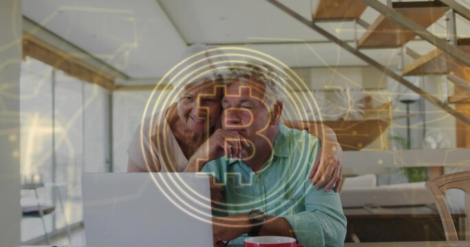 Leaning senior couple looking at laptop at home, viewing Bitcoin hologram, red mug, wristwatch