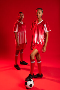 Female teammates posing in red studio, placing foot on black white soccer ball and wearing uniforms