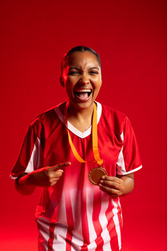 African American woman wearing red jersey holding gold medal and pointing, laughing in studio
