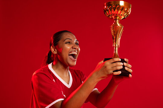 African woman holding gold trophy with black base, cheering in studio, red jersey, face paint