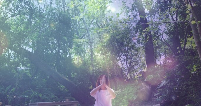 Standing woman wearing white T-shirt holding hands in forest glade with bench fallen trunk sunbeams