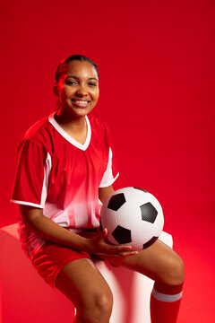 African American woman sitting on white cube in studio wearing red jersey holding soccer ball