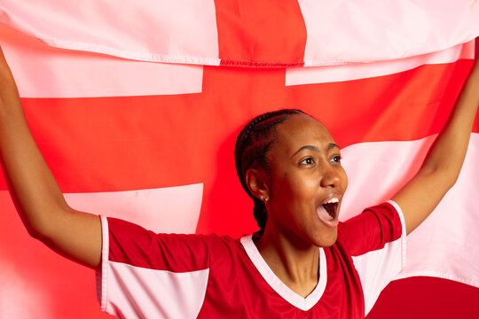 African woman holding red white cross flag aloft in studio, cheering, wearing red jersey with studs