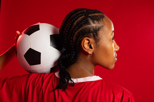 African American woman holding soccer ball on shoulder in studio with red backdrop, in red jersey