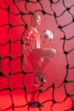 Woman wearing red soccer uniform juggling soccer ball behind goal net with haze in studio