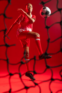 Female soccer player jumping, controlling ball in red kit through blurred black net on red backdrop